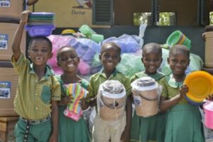 A group of five children in Ghana smiling and holding new kitchen supplies. They are wearing green school uniforms.